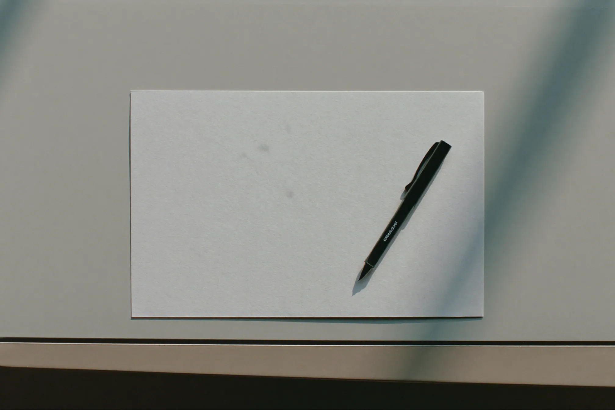 A single blank sheet of paper and a black fountain pen on a pale grey desk, with the shadow of a window frame across them